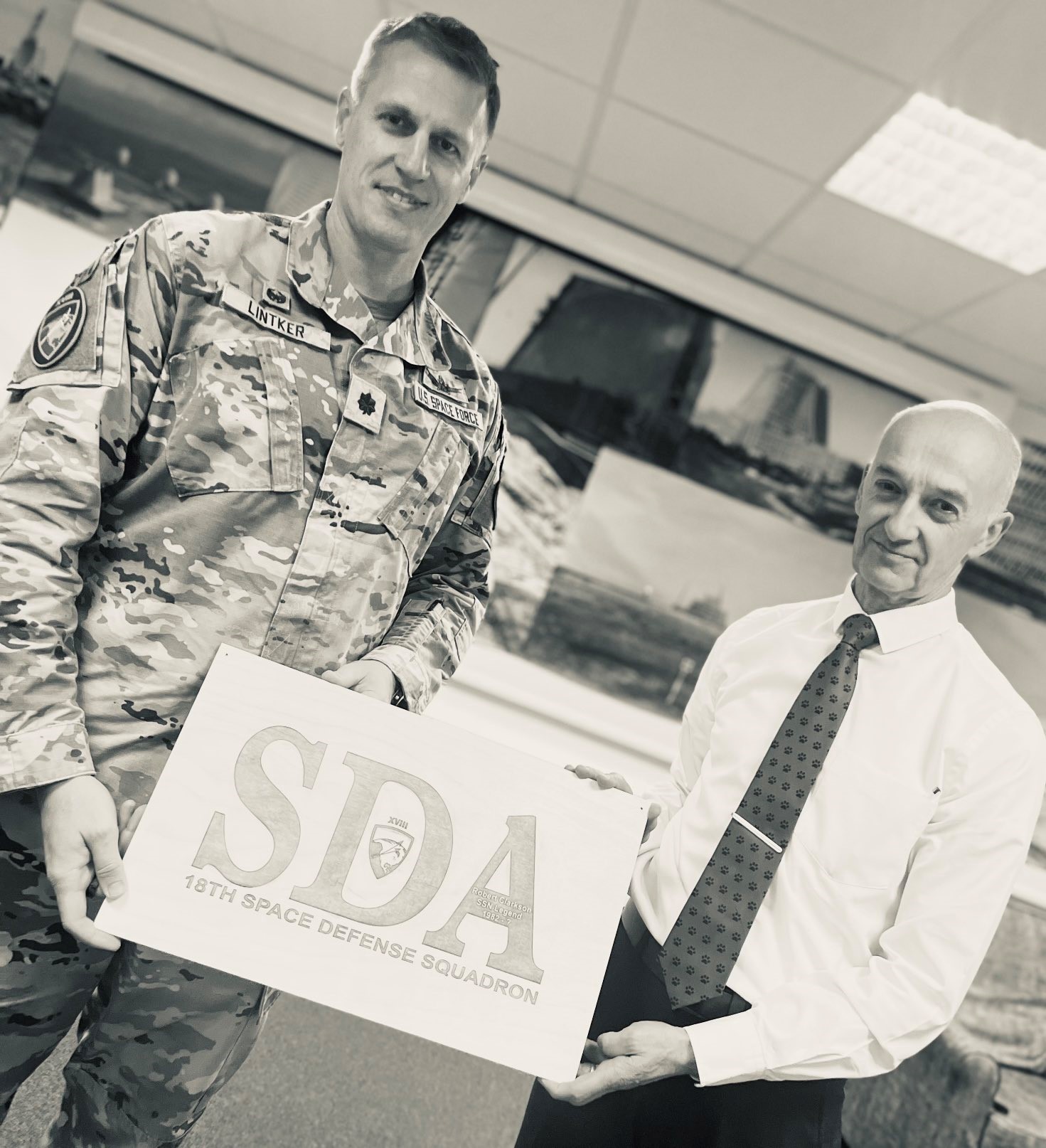 Black and white image shows RAF aviator and civilian holding a Space Defence Squadron sign. 